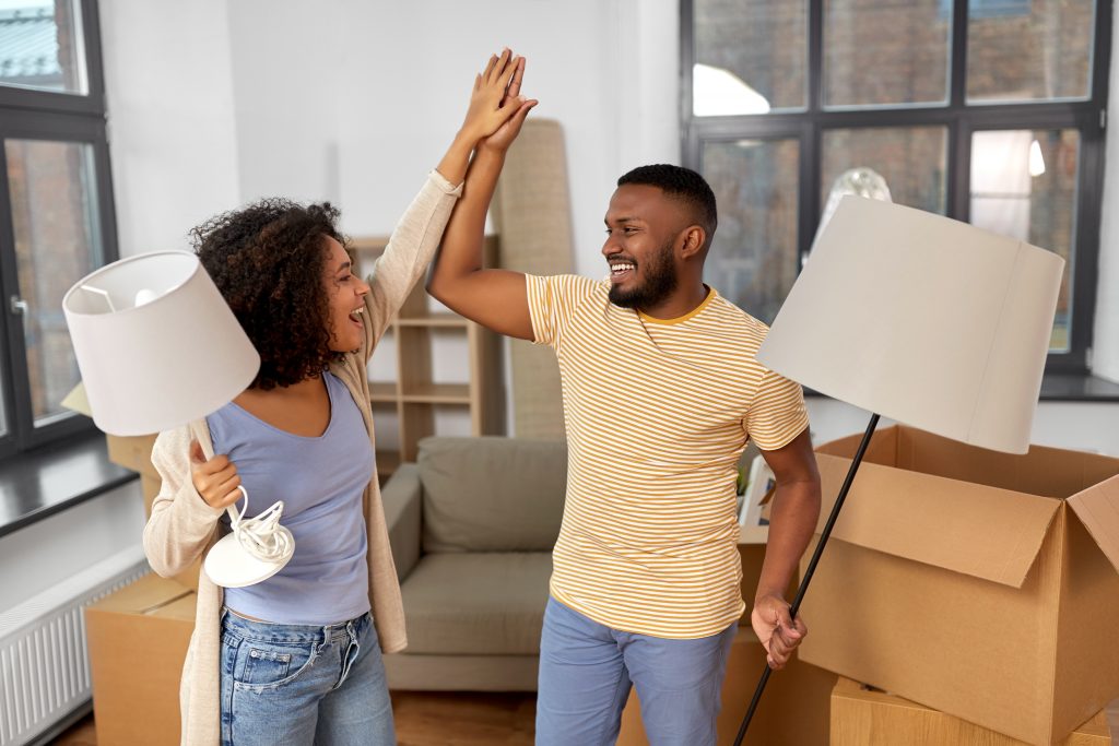 Een lachend Afro-Amerikaans koppel geeft elkaar een high-five terwijl ze lampen vasthouden, omringd door verhuisdozen.