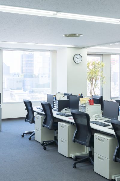 Een rij moderne bureaus met computers en bureaustoelen in een kantoor met grote ramen.