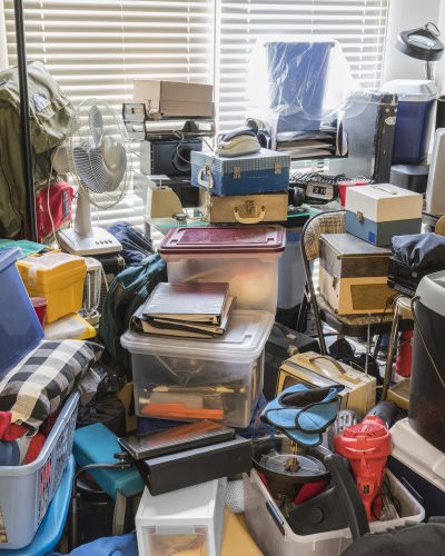 Een rommelige opslagruimte met stapels dozen, kleding, een ventilator en diverse huishoudelijke artikelen.